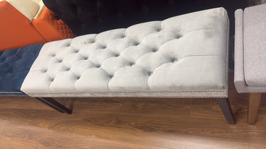 A light gray tufted velvet bench with dark metal legs, shown on a wooden floor next to other furniture.