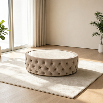 A round tufted ottoman or coffee table with a glass top sits on a light rug in a bright, modern room with wood floors, curtains, and potted plants.