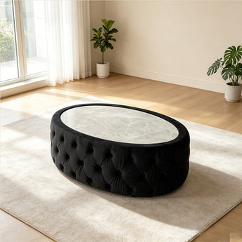 An oval tufted ottoman or coffee table with a black velvet upholstered base and a light marble-patterned top, shown in a bright modern room on a cream rug.