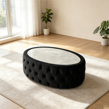 An oval tufted ottoman or coffee table with a black velvet upholstered base and a light marble-patterned top, shown in a bright modern room on a cream rug.