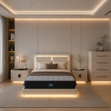 A modern bedroom featuring a low-profile platform bed with a tufted mattress, illuminated by warm LED under-bed lighting. The bed has a minimalist white headboard with integrated lighting, flanked by matching bedside tables, a white dresser, and a textured beige rug on a light wood floor.